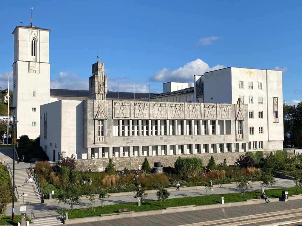 Bærum rådhus i ettermiddagssolen, omgitt av frodig grøntareal og en åpen plass hvor folk nyter den klare himmelen.